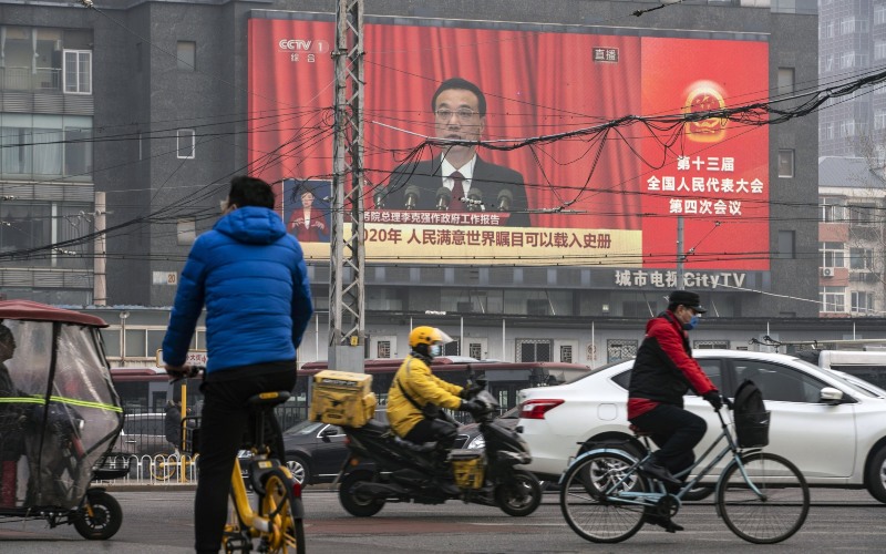 Para komuter melewati layar yang menunjukkan siaran langsung berita Perdana Menteri China Li Keqiang berbicara di Kongres Rakyat Nasional di Beijing, China, Jumat, (5/3 - 2021). Bloomberg / Qilai Shen